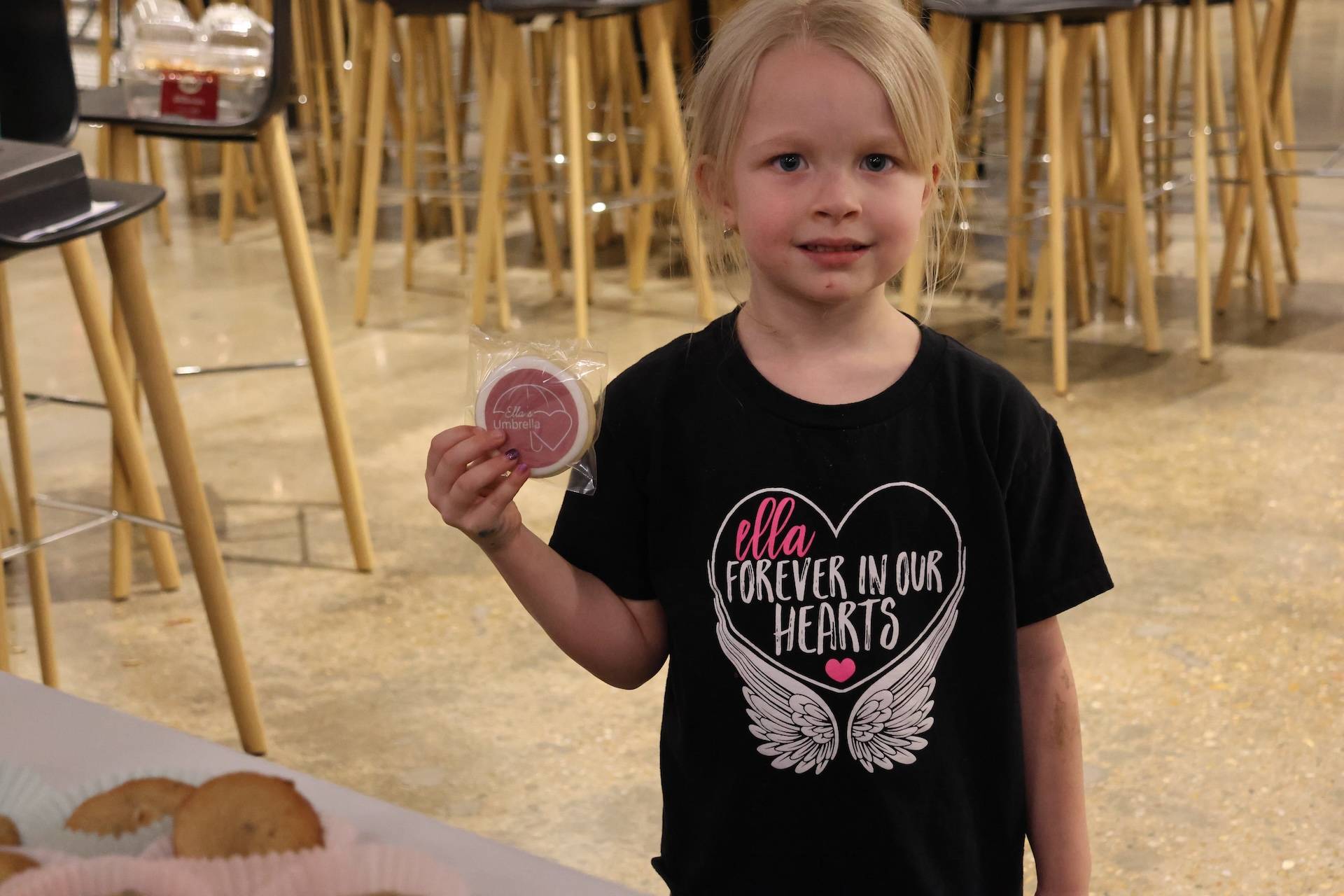 Young girl holding a wrapped cookie with a pink logo, wearing a black T-shirt that reads ella forever in our hearts, standing in a cafe-like room.
