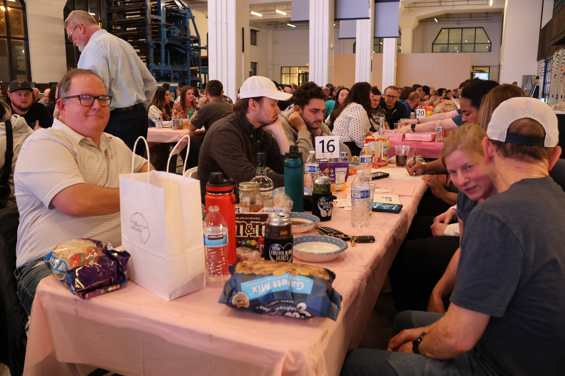Crowded indoor gathering with people seated at a long pink-covered table laden with snacks, drinks and a table number 16.