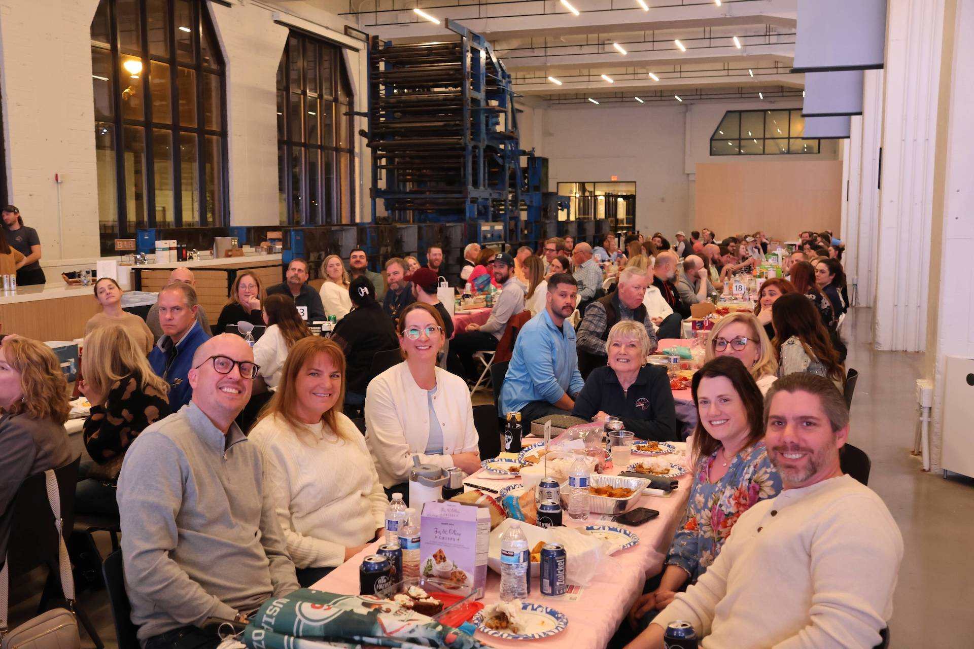 People seated at long banquet tables in a spacious industrial hall, smiling and sharing food.